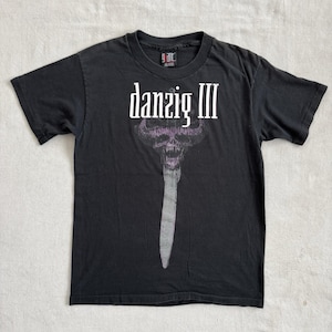 May include: Black t-shirt featuring the text "danzig III" in white above a purple and gray skull graphic with a long, gray blade. The shirt is a crew neck and appears to be made of cotton.