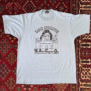 以下が含まれることがあります： ライトブルーの半袖Tシャツ。建物から顔を出す人物の黒いグラフィックプリントと、「RANCH APOCALYPSE」「We Ain't Comin' Out」「February 28, 1993」の文字がデザインされています。クルーネックで標準的なフィット感です。
