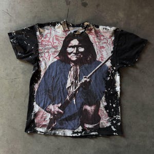 Op de afbeelding: Een vintage zwart-wit T-shirt met een vervaagde afbeelding van Geronimo, een Indiaanse leider, die een geweer vasthoudt. Het shirt heeft een kaart-achtig ontwerp met rode tekst en een zwart splatterpatroon.