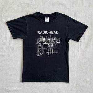 Puede incluir: Camiseta negra con el nombre de la banda "RADIOHEAD" en letras blancas en la parte superior de un gráfico blanco. El gráfico representa una figura, una cámara y un muro de ladrillos. La camiseta tiene mangas cortas y cuello redondo.