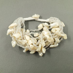 Könnte beinhalten: Ein weißes Armband mit unregelmäßigen, cremefarbenen Muschelteilen. Das Armband besteht aus mehreren Strängen kleiner, klarer Perlen und ist mit einer weißen Kordel zusammengebunden. Die Muschelteile sind im gesamten Armband verteilt.