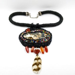 May include: A black beaded necklace with a large, oval pendant featuring a natural stone with brown, white, and black markings. The pendant is surrounded by colorful beads and has a small feather charm hanging from the bottom.