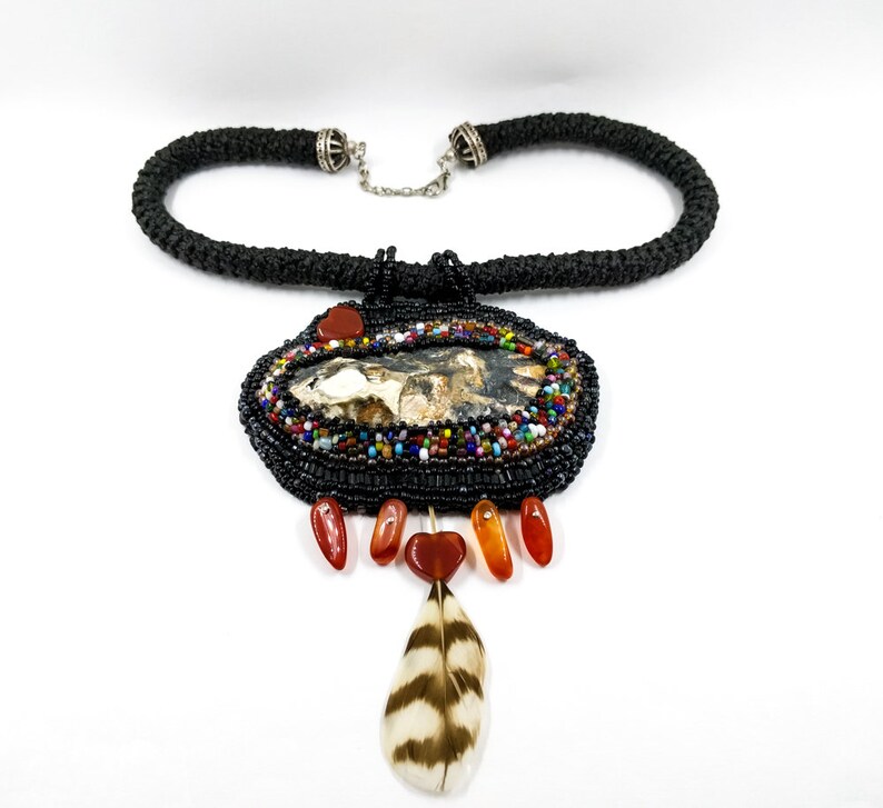 May include: A black beaded necklace with a large, oval pendant featuring a natural stone with brown, white, and black markings. The pendant is surrounded by colorful beads and has a small feather charm hanging from the bottom.