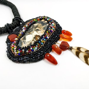 May include: A beaded necklace with a large, oval-shaped pendant featuring a natural stone with brown and white markings. The pendant is surrounded by black beads and accented with colorful beads. The necklace also features a brown and white feather charm and several orange-red gemstone accents.