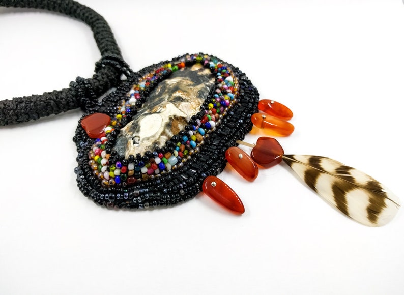 May include: A beaded necklace with a large, oval-shaped pendant featuring a natural stone with brown and white markings. The pendant is surrounded by black beads and accented with colorful beads. The necklace also features a brown and white feather charm and several orange-red gemstone accents.