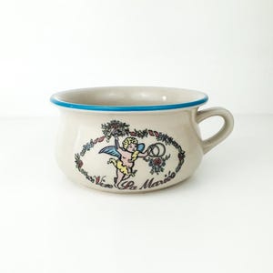 Può includere: Una zuppiera in ceramica bianca con un bordo blu e un disegno floreale con un cherubino che tiene degli anelli nuziali. La zuppiera ha il testo "Vive La Marie" sul fondo.