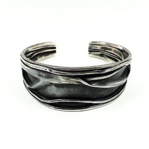 May include: A silver cuff bracelet with a textured, wavy design. The bracelet has a dark, oxidised finish and an open design. The metal appears layered, creating a unique, artistic look. The bracelet is shown against a white background.
