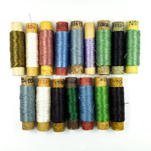 May include: A collection of 18 spools of thread in various colors, including green, blue, red, black, white, and brown. The spools are wrapped in paper and have labels with numbers and letters.