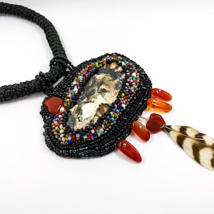May include: A beaded necklace with a large, oval pendant featuring a natural stone and a feather charm. The pendant is surrounded by colorful beads and has a black cord.