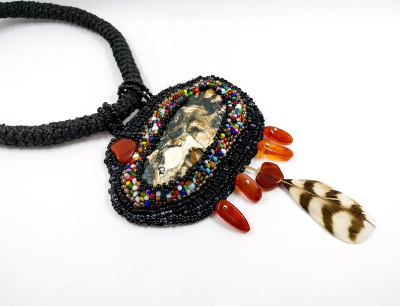 May include: A beaded necklace with a large, oval pendant featuring a natural stone and a feather charm. The pendant is surrounded by colorful beads and has a black cord.