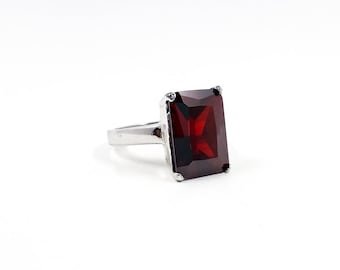 May include: A silver ring with a large, rectangular, deep red gemstone. The gemstone is the focal point, set in a simple silver band. The ring is displayed against a white background, highlighting its colour and design.
