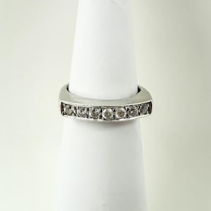 Puede incluir: Un anillo plateado con una fila de pequeñas piedras redondas y claras. El anillo se muestra sobre un objeto cilíndrico blanco, resaltando su diseño. Las piedras están espaciadas uniformemente.