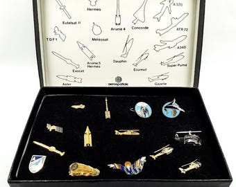 Op de afbeelding: Een zwarte doos met een verzameling kleine, gedetailleerde spelden en badges. De spelden tonen verschillende vliegtuigen en ruimtevaartuigen, sommige met kleurrijke emaille ontwerpen. Het deksel van de doos heeft lijntekeningen van vliegtuigen met labels zoals "Concorde" en "Ariane 4".