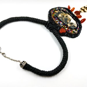 May include: A black beaded necklace with a large, oval pendant featuring a brown and white agate stone surrounded by colorful beads and a small feather. The pendant is accented with red and orange beads.