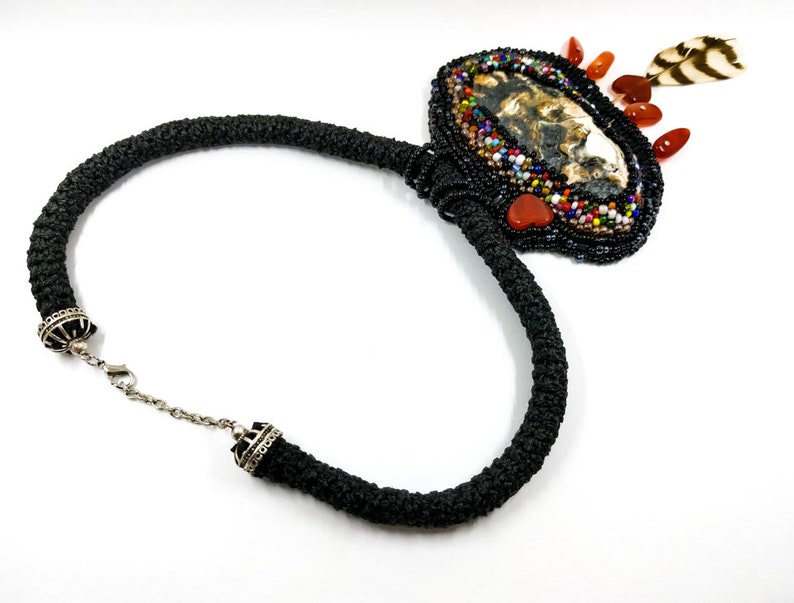 May include: A black beaded necklace with a large, oval pendant featuring a brown and white agate stone surrounded by colorful beads and a small feather. The pendant is accented with red and orange beads.