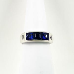 Puede incluir: Un anillo de banda en tono plateado con tres piedras de color azul intenso de corte cuadrado engastadas horizontalmente en el centro, flanqueadas por una pequeña piedra redonda transparente a cada lado, con un diseño simétrico y acabado pulido.