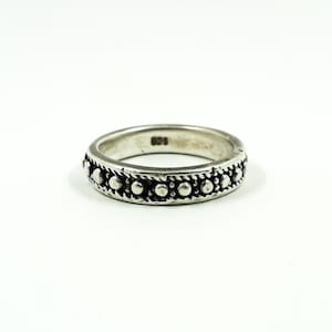 Puede incluir: Un anillo de plata con un diseño de cuentas. El anillo presenta una serie de pequeñas cuentas en relieve alrededor de la banda, intercaladas con una textura similar a una cuerda. El anillo está sobre un fondo blanco.