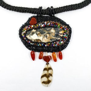 May include: A beaded necklace with a large oval pendant featuring a natural stone, surrounded by colorful beads. The pendant is accented with red gemstone accents and a brown and white feather.