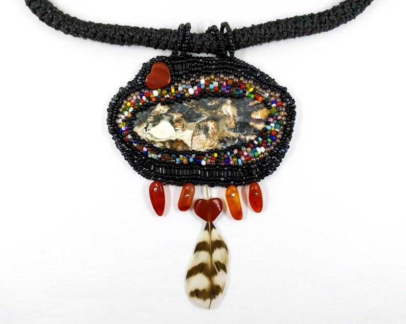 May include: A beaded necklace with a large oval pendant featuring a natural stone, surrounded by colorful beads. The pendant is accented with red gemstone accents and a brown and white feather.