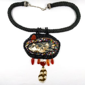 May include: A black beaded necklace with a large, oval pendant featuring a natural stone, surrounded by colorful beads. The pendant is accented with red beads and a feather.