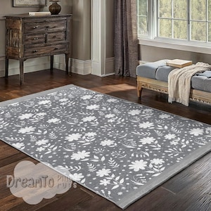 Puede incluir: Una alfombra gris con un patrón floral blanco. La alfombra tiene un borde gris y se muestra sobre un suelo de madera.