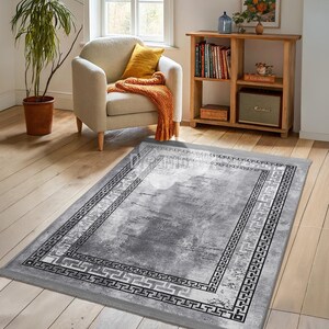 Könnte beinhalten: Ein rechteckiger grauer Teppich mit einem Used-Look-Muster und einem geometrischen Rand. Der Teppich hat ein hellgraues Mitteldesign mit dunkleren grauen Akzenten und einem schwarz-weißen Rand. Der Teppich liegt auf einem Holzboden.