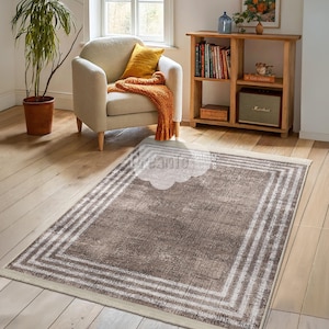 Peut inclure: Un tapis de salon brun et crème avec une bordure blanche et un aspect vieilli. Le tapis est posé sur un sol en bois dans un salon.