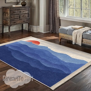 May include: A rectangular area rug with a mountain landscape design. The rug features shades of blue, with a red sun in the upper portion. The rug has a cream-colored border and fringe. The text "DreamTo Pillows Rugs" is visible in the lower left corner.