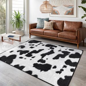 May include: A black and white cowhide patterned area rug with a white background and black spots. The rug is laid out on a wooden floor in a living room setting with a brown leather sofa and a coffee table.
