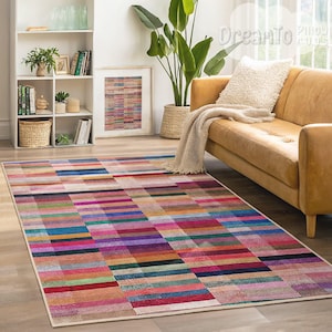 Puede incluir: Una alfombra rectangular colorida con un patrón geométrico de rayas horizontales y verticales en tonos de rosa, morado, azul, verde, rojo y marrón. La alfombra está colocada sobre un suelo de madera en un salón.