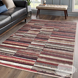 Puede incluir: Una alfombra de área multicolor con un patrón a rayas en tonos de rojo, marrón, beige y blanco. La alfombra tiene un borde de color crema.