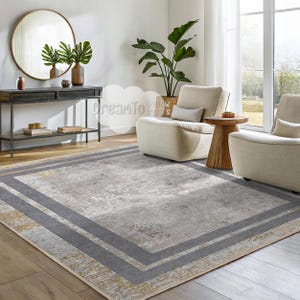 Op de afbeelding: Een rechthoekig vloerkleed met een verweerd, gedempt grijs en goud ontwerp. Het vloerkleed heeft een donkergrijze rand met een lichter grijs interieur. Het vloerkleed staat in een woonkamer met meubels en decor in neutrale tinten.