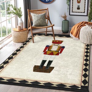 Puede incluir: Una alfombra rectangular con un fondo color crema y un borde de rombos negros y rojos. La alfombra presenta una gran ilustración de un soldado cascanueces en rojo, amarillo y negro. La alfombra está colocada en una habitación con una silla y una ventana.