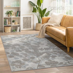 Puede incluir: Una alfombra rectangular con un diseño botánico en tonos grises y azules. La alfombra está colocada sobre un suelo de madera en un salón, con un sofá de cuero marrón y una estantería blanca al fondo.