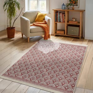 May include: A rectangular area rug with a repeating fan-shaped pattern in red and cream. The rug has a cream border with a geometric design. The rug is placed on a hardwood floor in a living room setting.