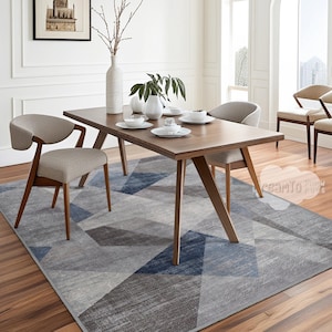 May include: A wooden dining table with four chairs sits on a grey and blue geometric area rug. The table has a light wood finish and the chairs are upholstered in a light grey fabric.