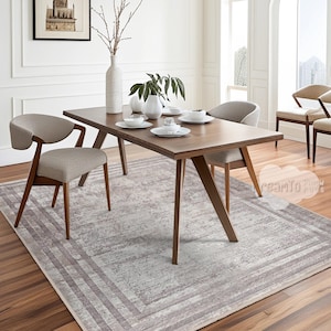 May include: A light brown wooden dining table with four chairs, two of which are visible in the image. The table is set with white plates, bowls, and cups. A light gray area rug with a patterned border is under the table.