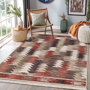 Op de afbeelding: Een rechthoekig vloerkleed met een geometrisch patroon in tinten bruin, beige en rood. Het vloerkleed heeft een rand en een herhalend ontwerp van gestileerde driehoekige vormen. Het vloerkleed ligt op een houten vloer in een kamer met natuurlijk licht.