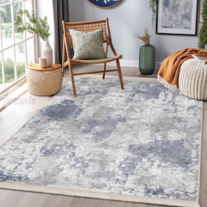 May include: A light gray and blue abstract patterned area rug with a cream-colored border. The rug is in a living room setting with a wooden chair and a side table.