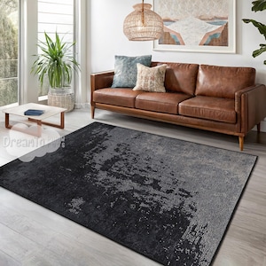 Può includere: Un tappeto da soggiorno astratto nero e grigio con un aspetto usurato e testurizzato. Il tappeto è posizionato in un ambiente da salotto con un divano in pelle marrone e un tavolino da caffè.