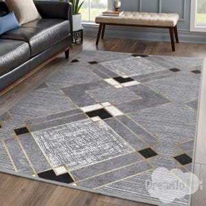 Puede incluir: Una alfombra geométrica gris y blanca con detalles dorados. La alfombra tiene un diseño moderno con cuadrados negros y blancos y un centro blanco desgastado.