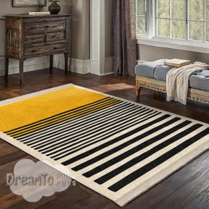 May include: A rectangular rug with a contemporary design. The rug has a yellow upper section, divided by black lines from the lower section with black and white stripes. The rug is on a dark wood floor.