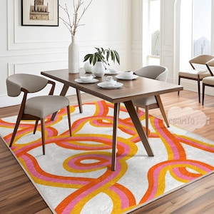 Può includere: Una sala da pranzo con un tavolo in legno, quattro sedie e un tappeto fantasia. Il tappeto ha una base bianca con linee curve arancioni, rosa e gialle. Le sedie hanno sedute in tessuto grigio e telai in legno. Il tavolo è lungo circa 180 cm.