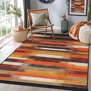 Pode incluir: Um tapete de área listrado colorido com uma borda preta. O tapete tem um padrão geométrico com listras de laranja, vermelho, marrom, bege e cinza. O tapete está colocado em um piso de madeira em uma sala de estar.