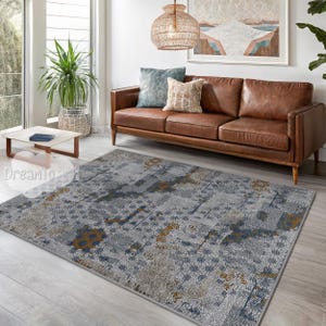 May include: A gray, blue, and gold patterned area rug with a geometric design. The rug is in a living room setting with a brown leather sofa and a potted plant.