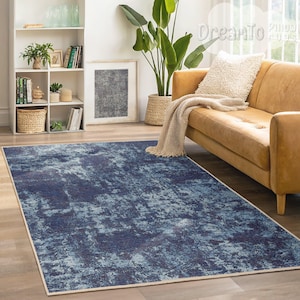 May include: A rectangular area rug with a distressed, abstract design in shades of blue and white. The rug has a cream-colored border and is placed on a wooden floor. The rug is in a living room setting with a leather sofa and a bookshelf.
