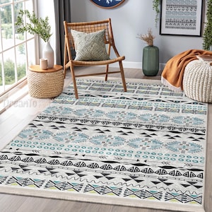 May include: A white and blue patterned area rug with geometric designs. The rug has a fringed border and is laid out on a wooden floor.