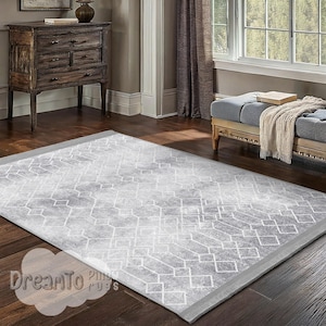 Puede incluir: Una alfombra de área gris y blanca con un patrón geométrico y un aspecto desgastado sutil. La alfombra tiene un borde gris claro.