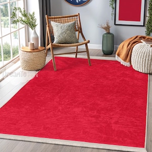 May include: A bright red rectangular rug with a textured surface. The rug has a cream border and is placed on a wooden floor. The rug is the focal point of a living room setting, with a chair and side table visible.