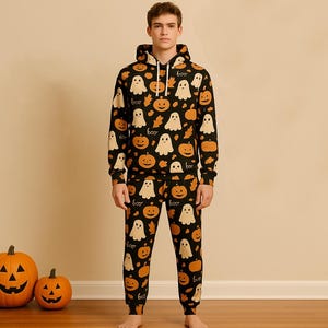 Puede incluir: Un conjunto de pijama negro con temática de Halloween con un patrón repetido de fantasmas blancos, calabazas naranjas y la palabra "boo". El conjunto incluye una sudadera con capucha y pantalones a juego. Dos calabazas talladas están en el suelo.